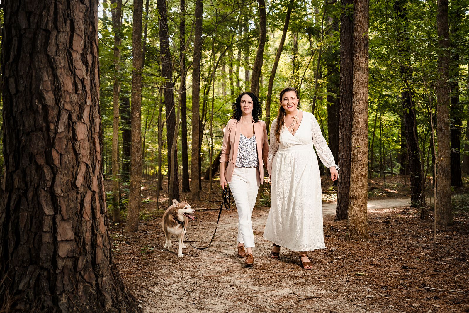 Two women walk hand in hand through the woods with their dog. It's their wedding day. 