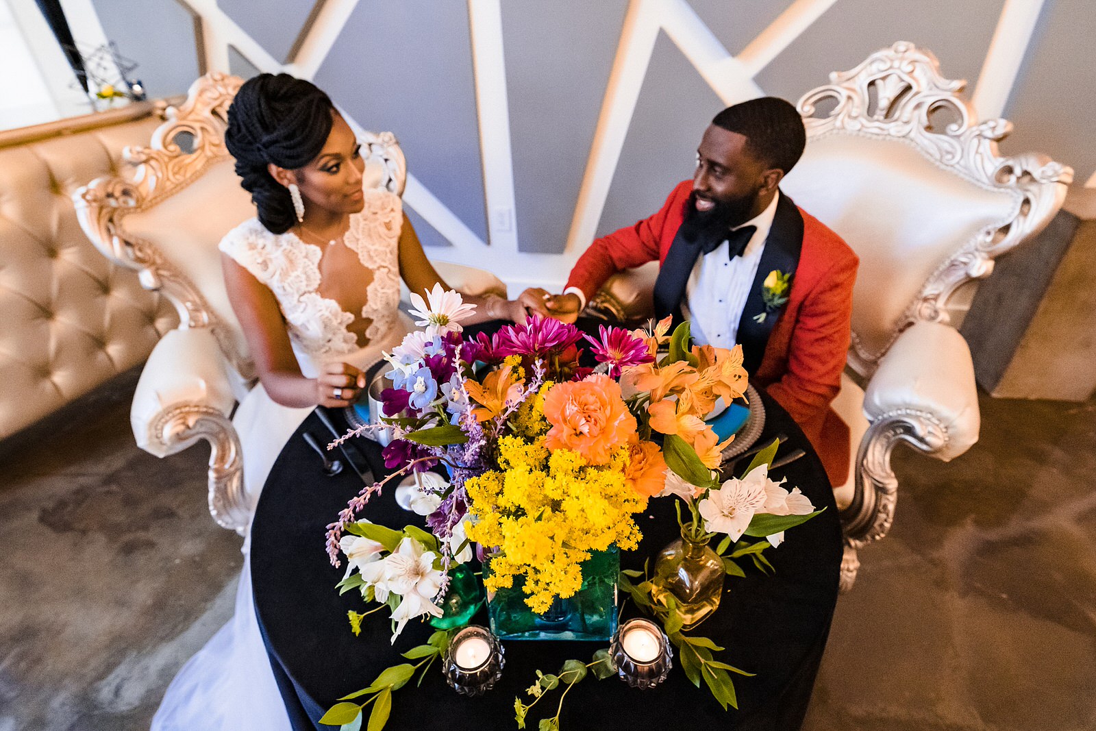 Couple sits at sweetheart table during wedding styled shoot. A large colorful bouquet sits in front of them.