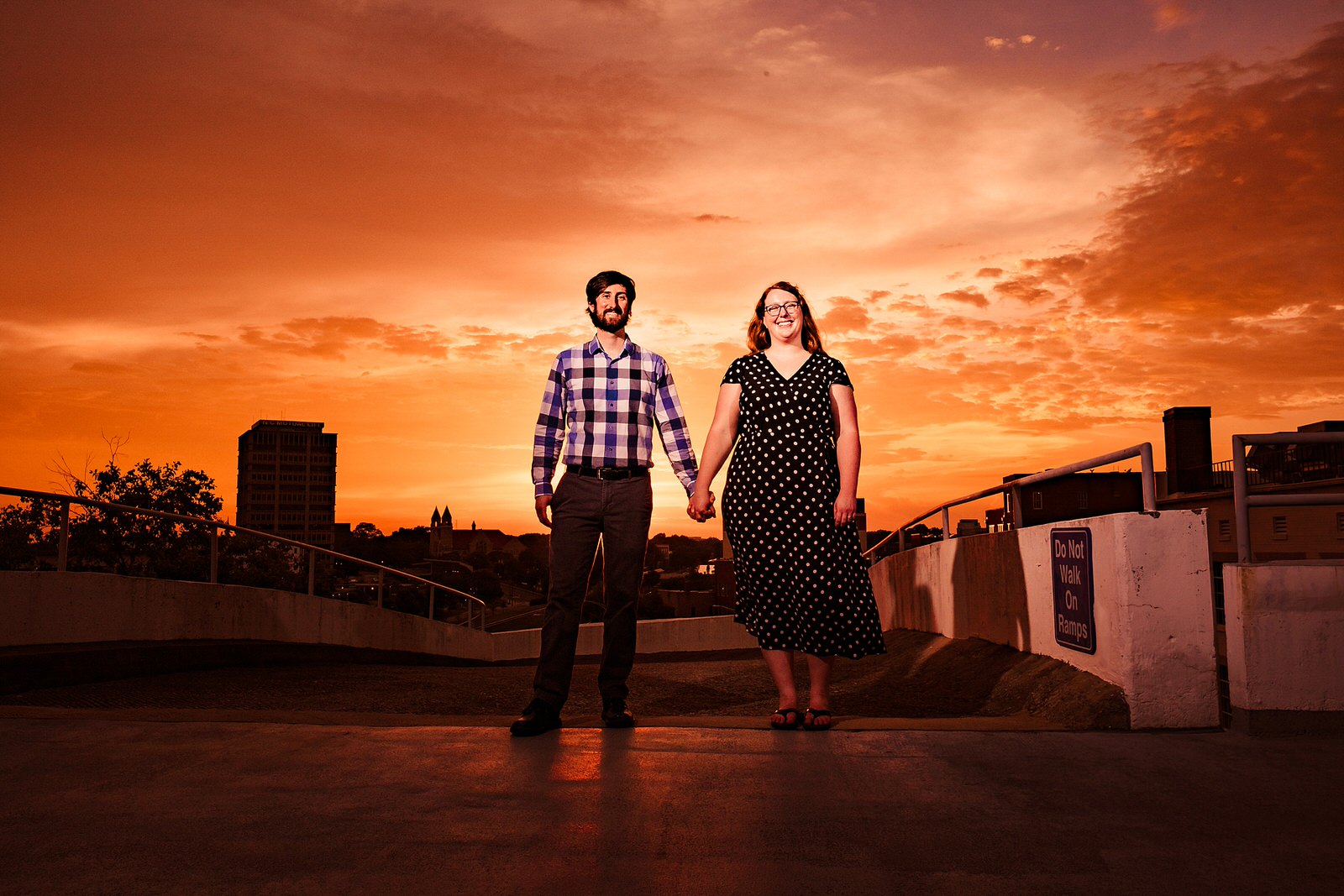 An engaged couple holds hands in front of an orange and blue sunset