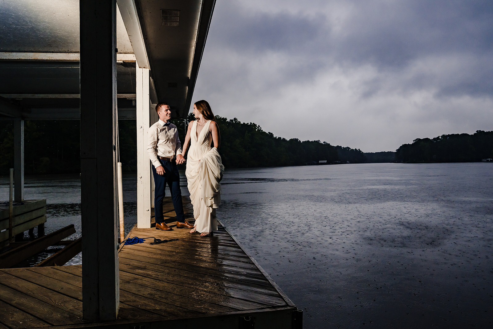bride and groom stand on a covered boat dock as rain pours over the lake