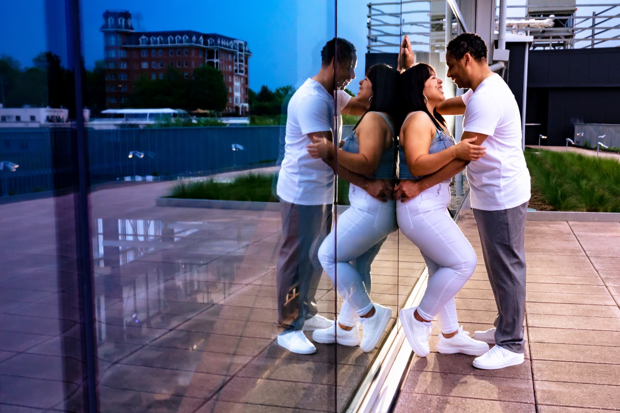 couple leans against a floor to ceiling window on an outdoor patio, the city of Raleigh at blue hour is reflected in the glass to the left of the couple