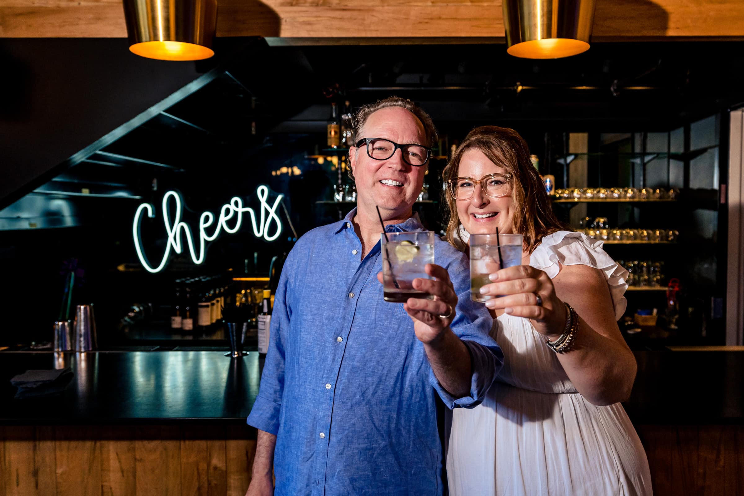 couple in casual wedding attire stands in front of a bar with a neon sign that says "Cheers" and the couple is holding their drinks out toward the camera | photo by Kivus & Camera