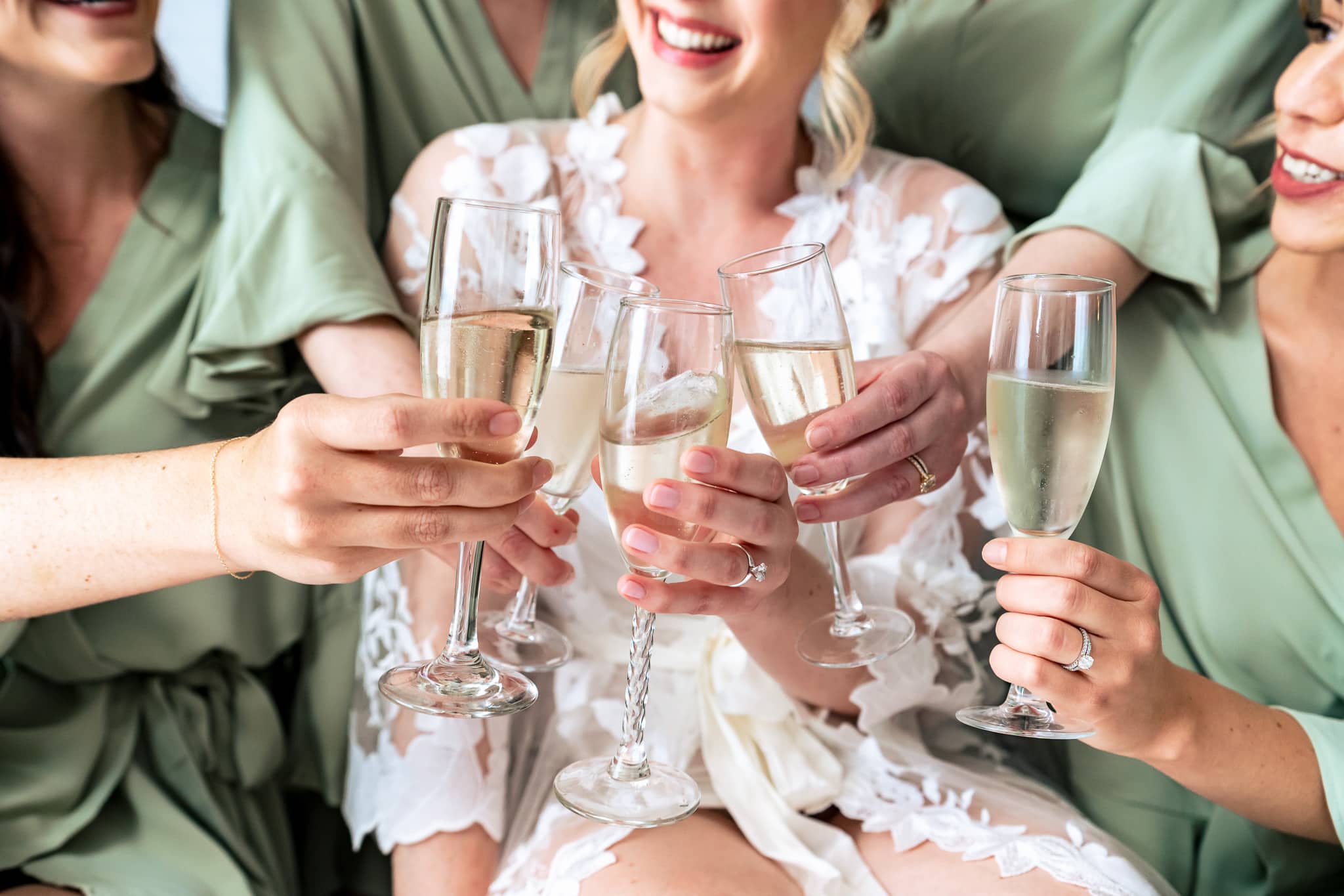 closeup of champagne glasses toasting, glasses are held by a bride in a white lace robe and bridesmaids in sage green silk robes | photo by Kivus & Camera