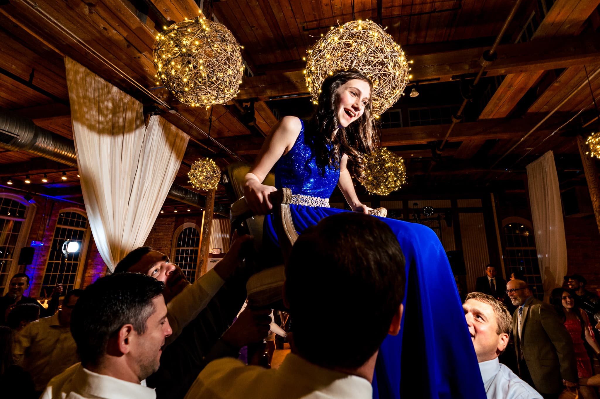 young woman in a blue dress is lifted in a chair during the hora at her bat mitzvah reception
