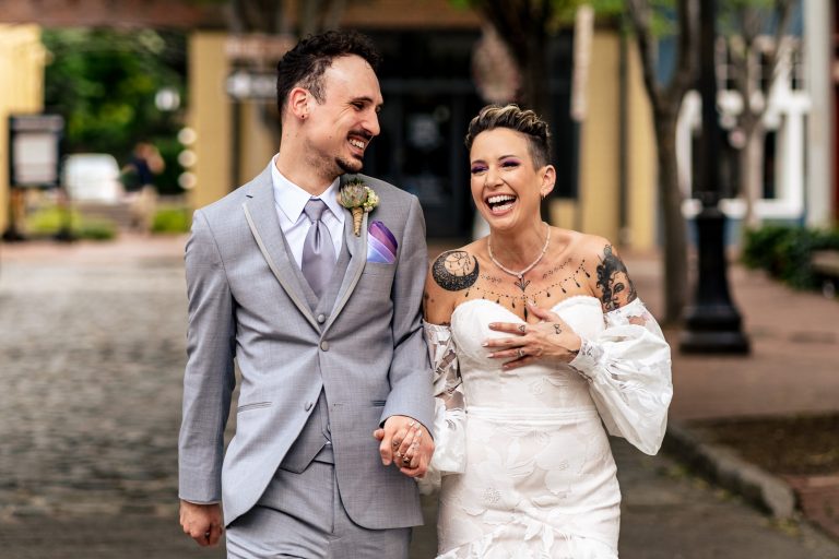 couple in wedding attire laughs and walks hand in hand down a cobblestone street at the Grove at City Market in Raleigh, NC