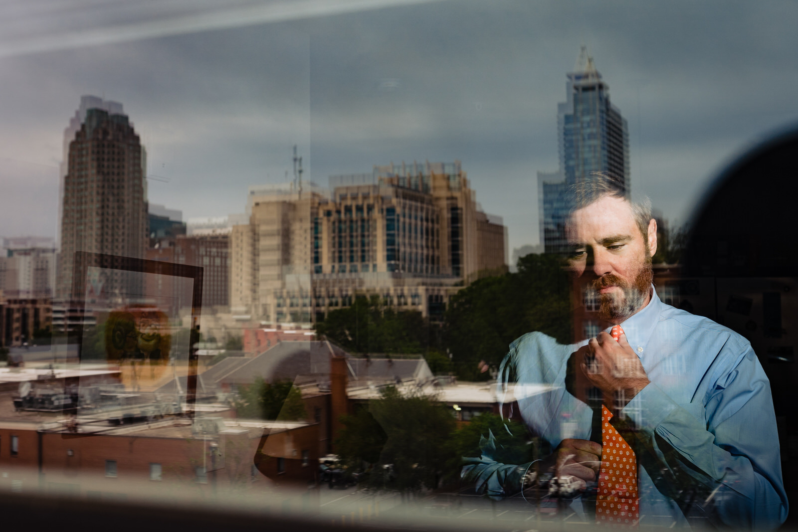 photo of groom tying his tie, with donwtown Raleigh reflected in the window