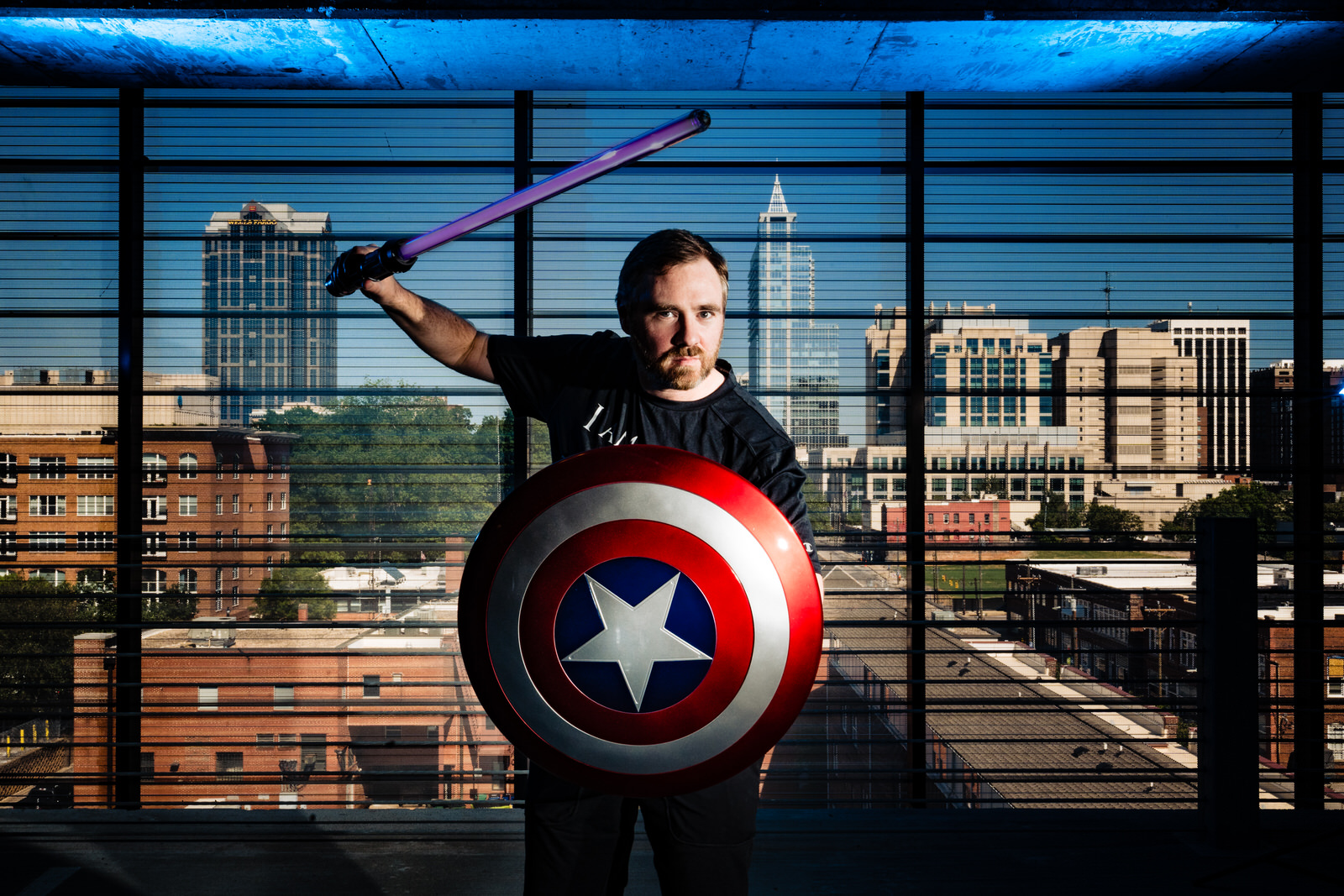 nerdy wedding photographer poses with light saber, captain america shield, and Raleigh skyline