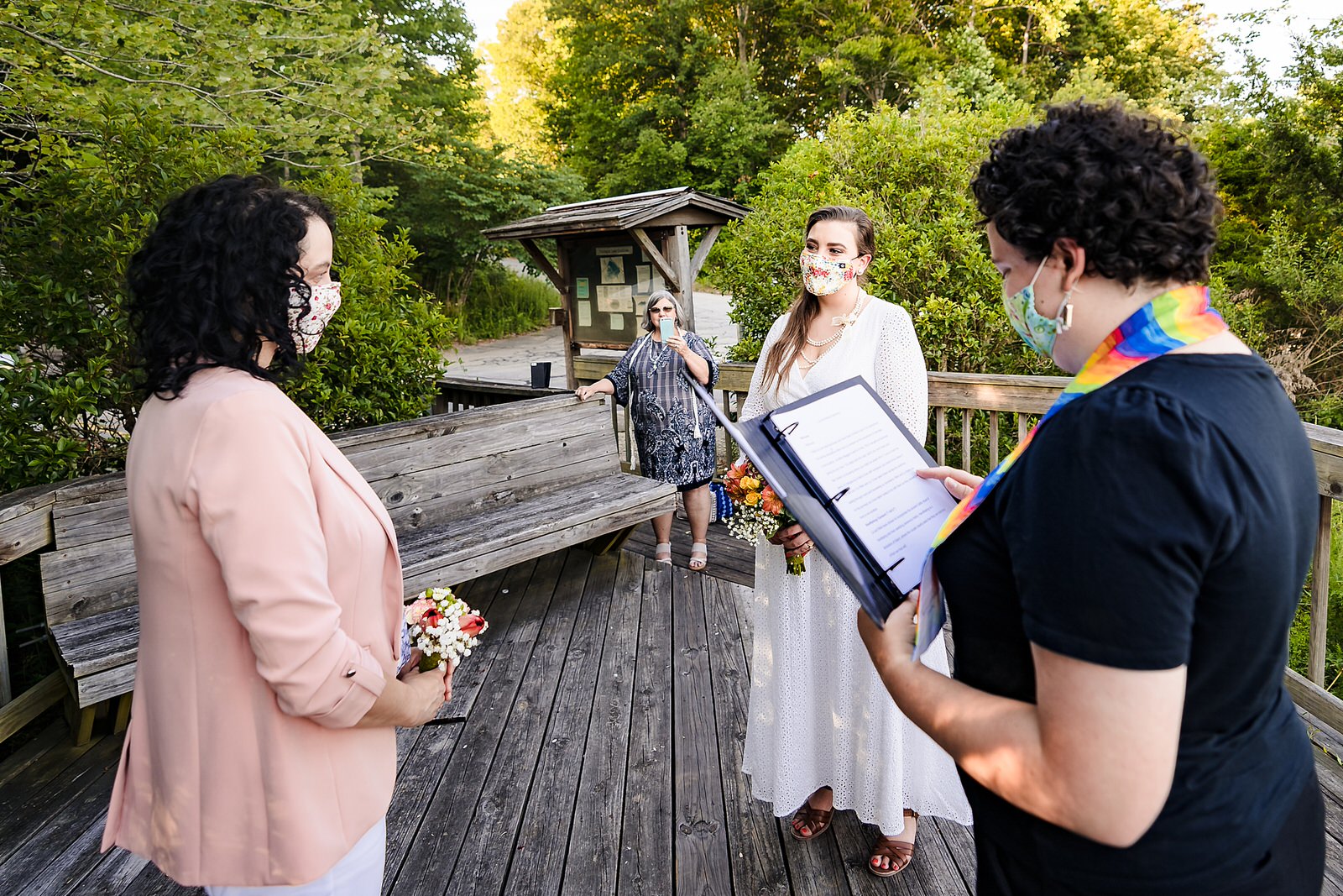 Two brides stand in front of a wedding officiant. All three wear masks to help prevent the spread of covid19. 