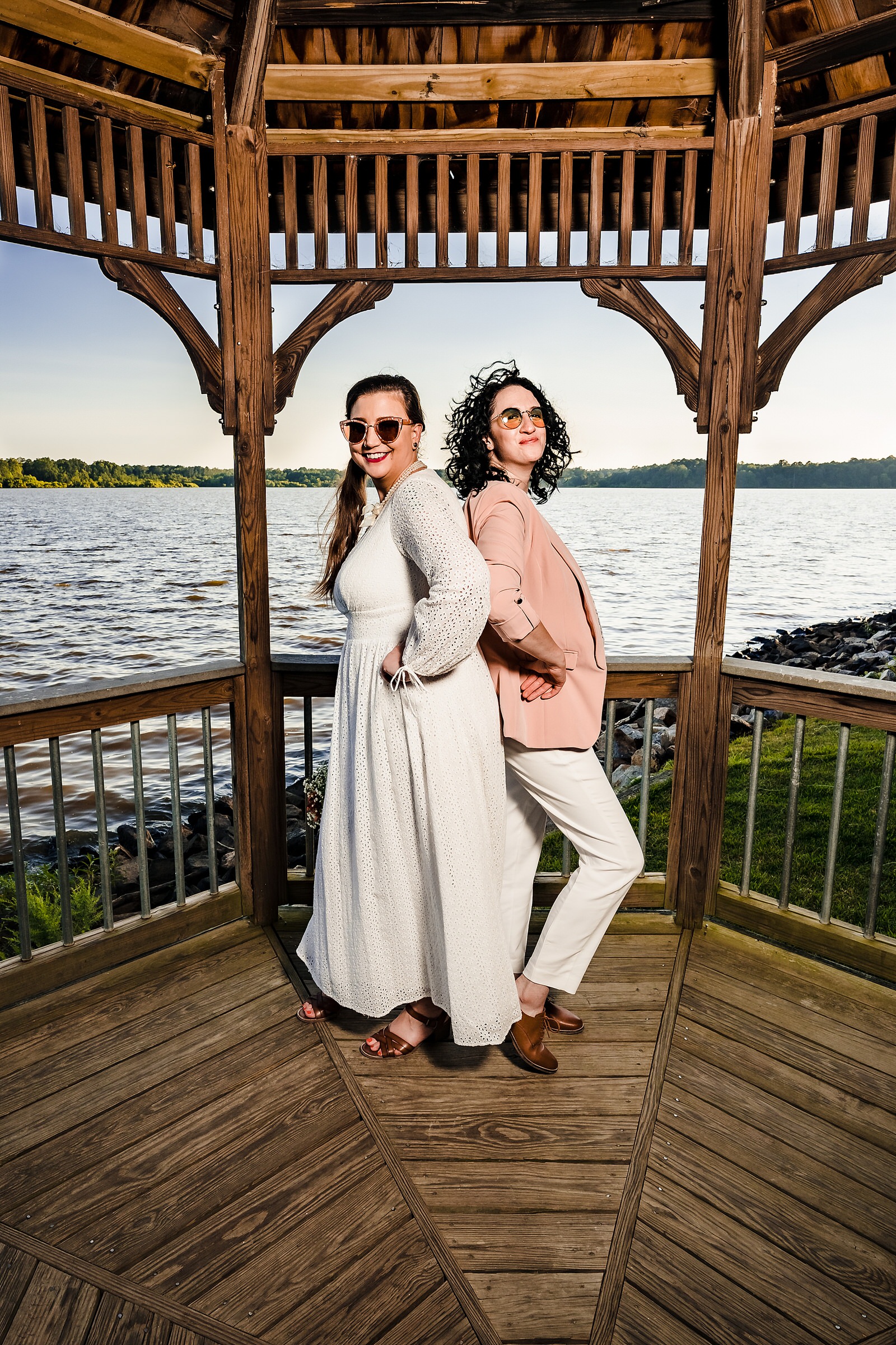 Two brides pose in fashionable outfits and sunglasses on their wedding day. 