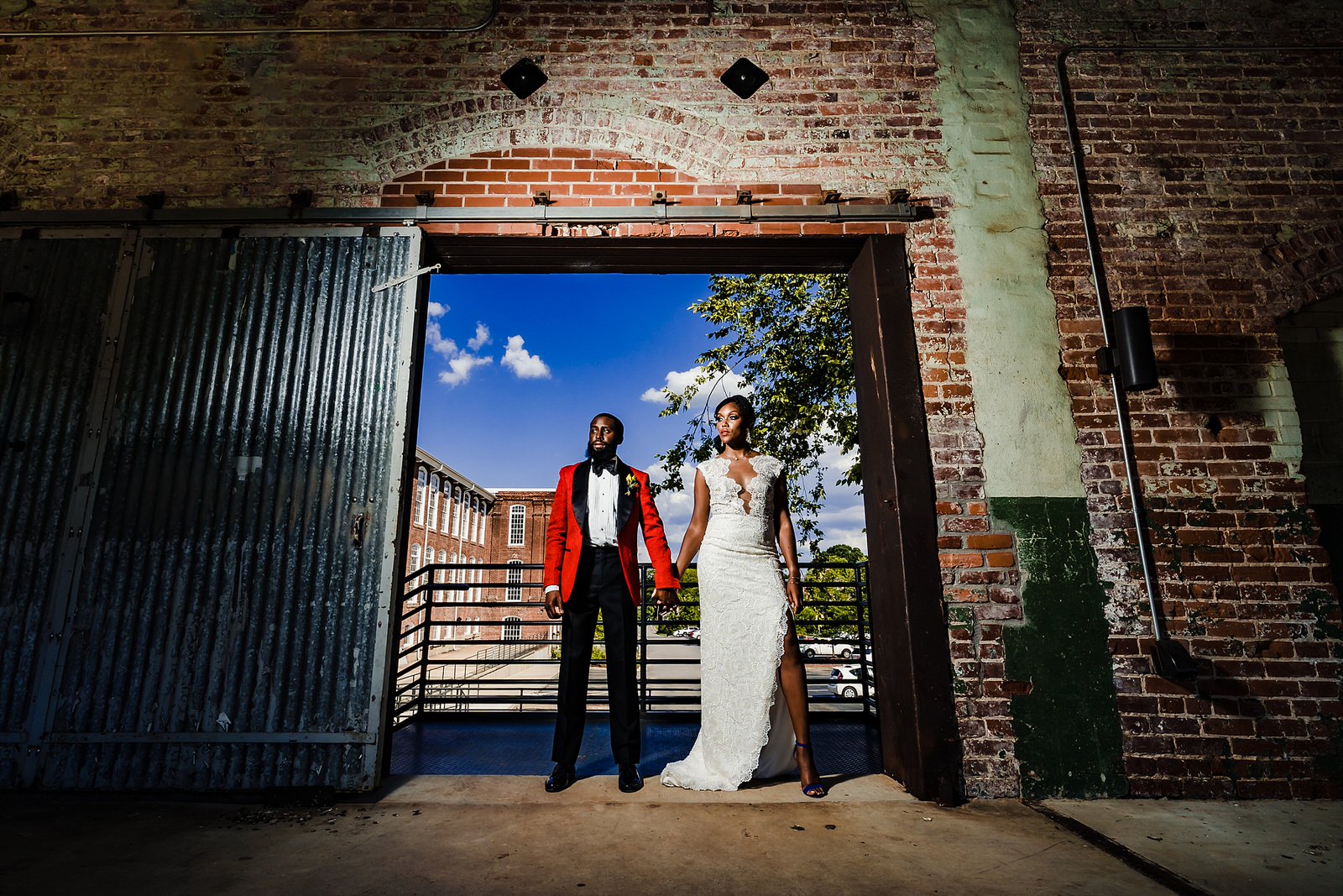 A Black couple stands in wedding gown and custom red suit at Belt Line Station in Durham, NC