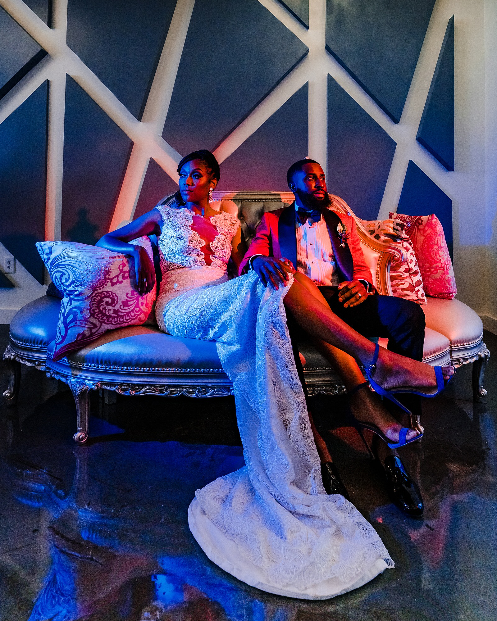 Dramatically lit portrait of a bride and groom sitting on a couch. The groom is lit with red light and the bride is lit with blue light. 