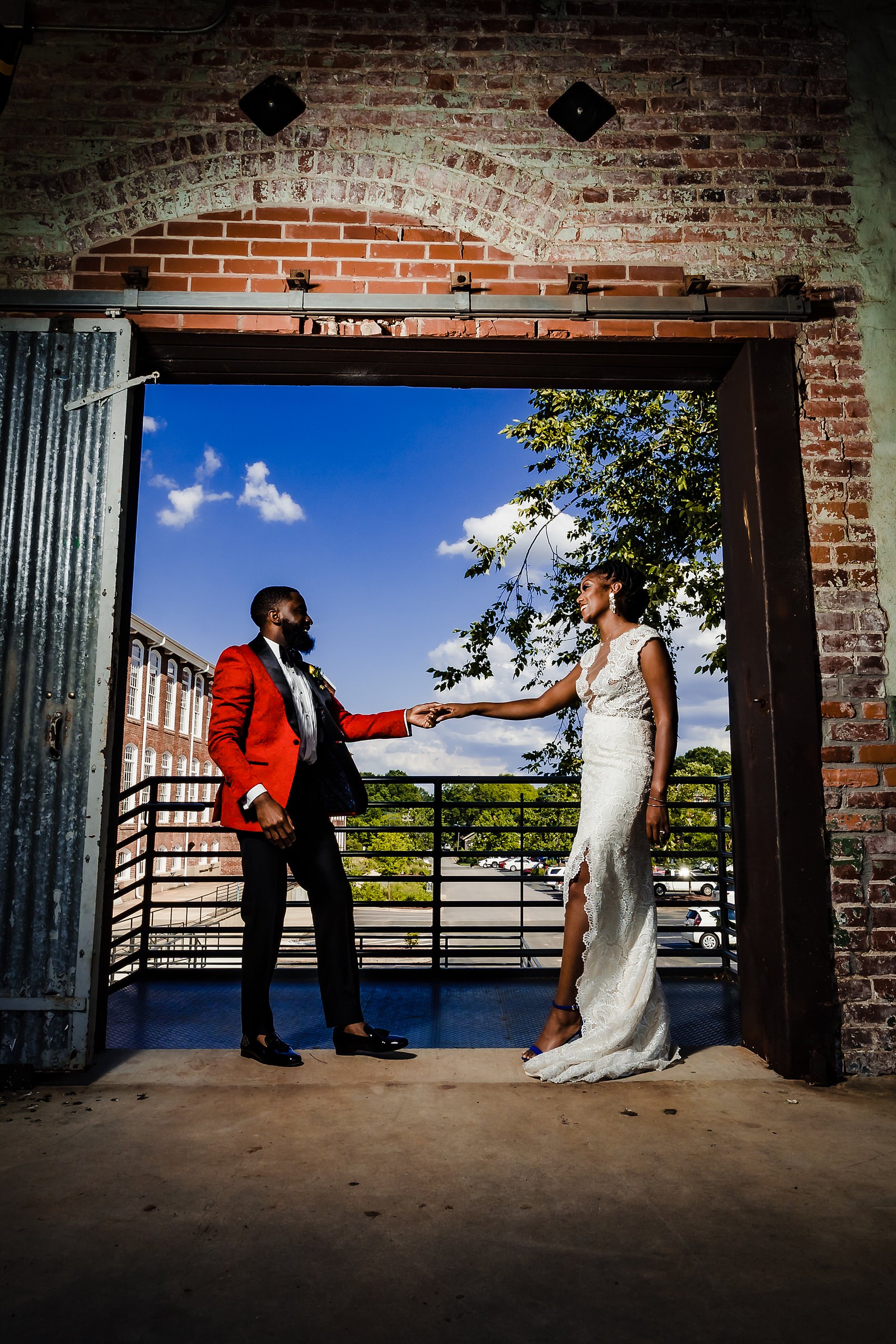 A Black couple laughs and dances in wedding gown and custom red suit at Belt Line Station in Durham, NC