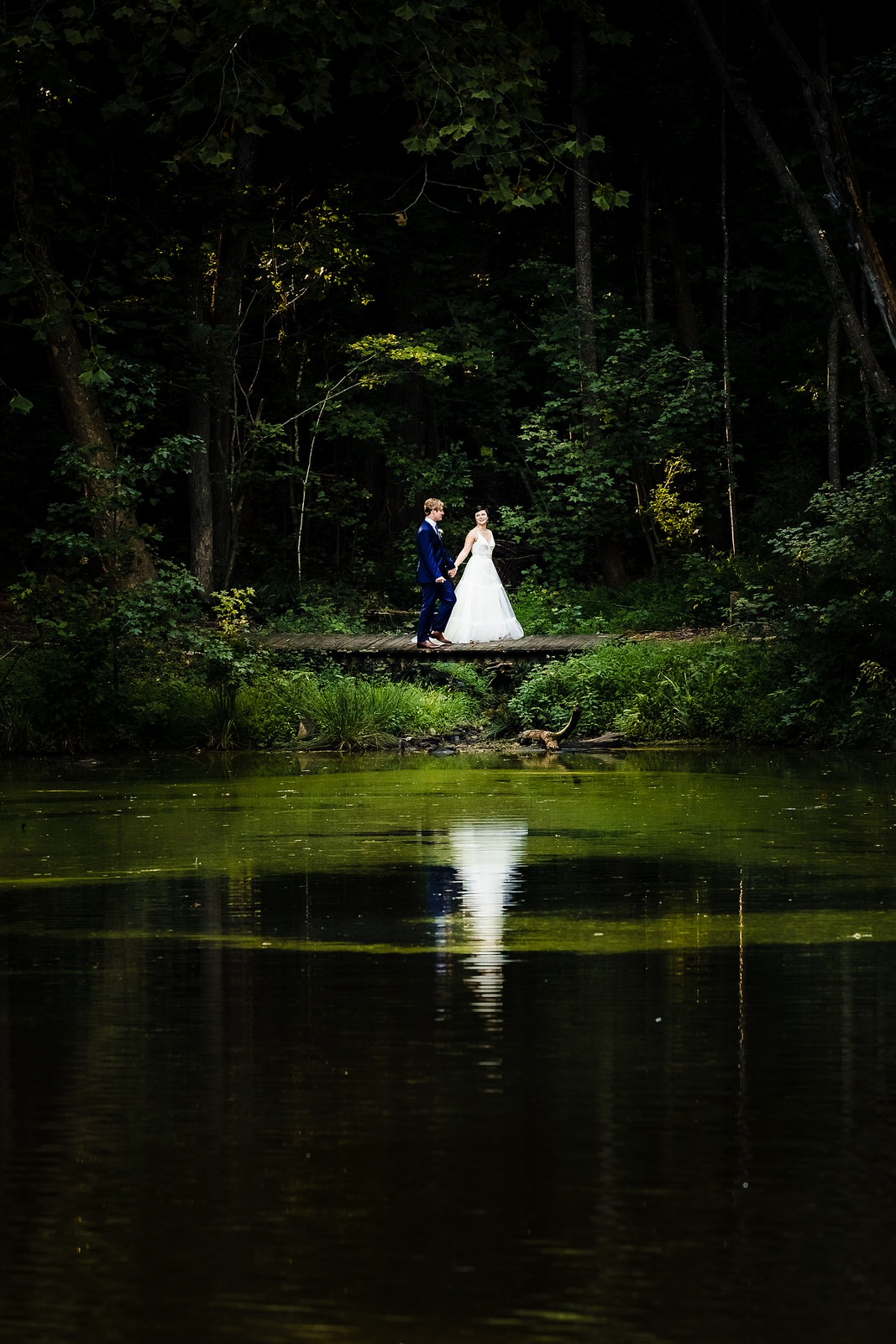 bride and groom walk along a lakeside, they are reflected in the lake's surface