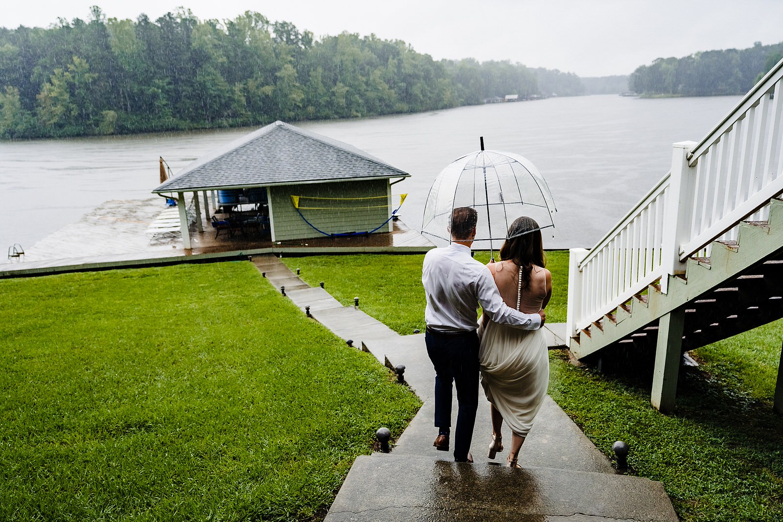 groom escorts bride down the path to a lake in the pouring rain