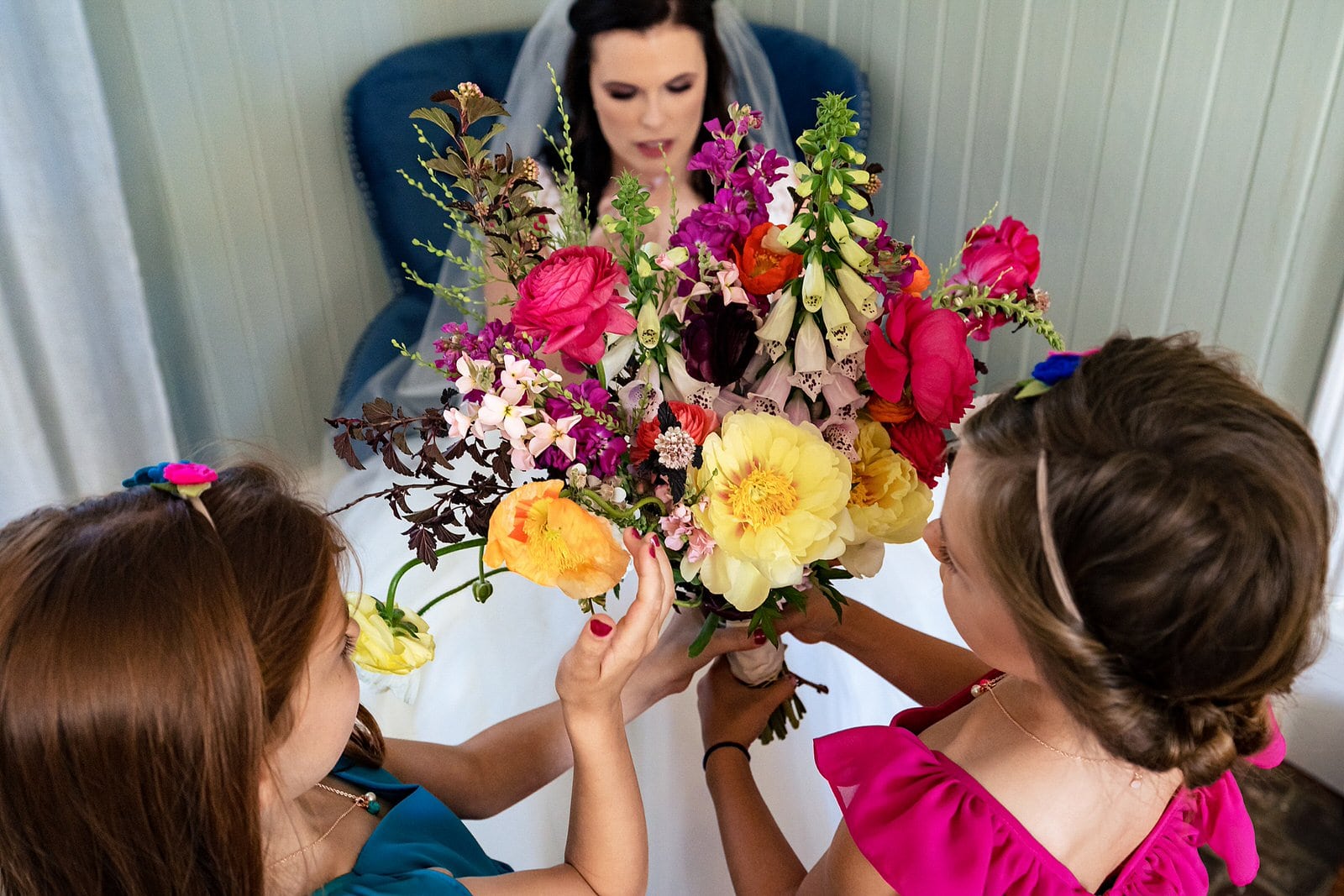 Flower girls hold bride's colorful bouquet as she reapplies lip gloss
