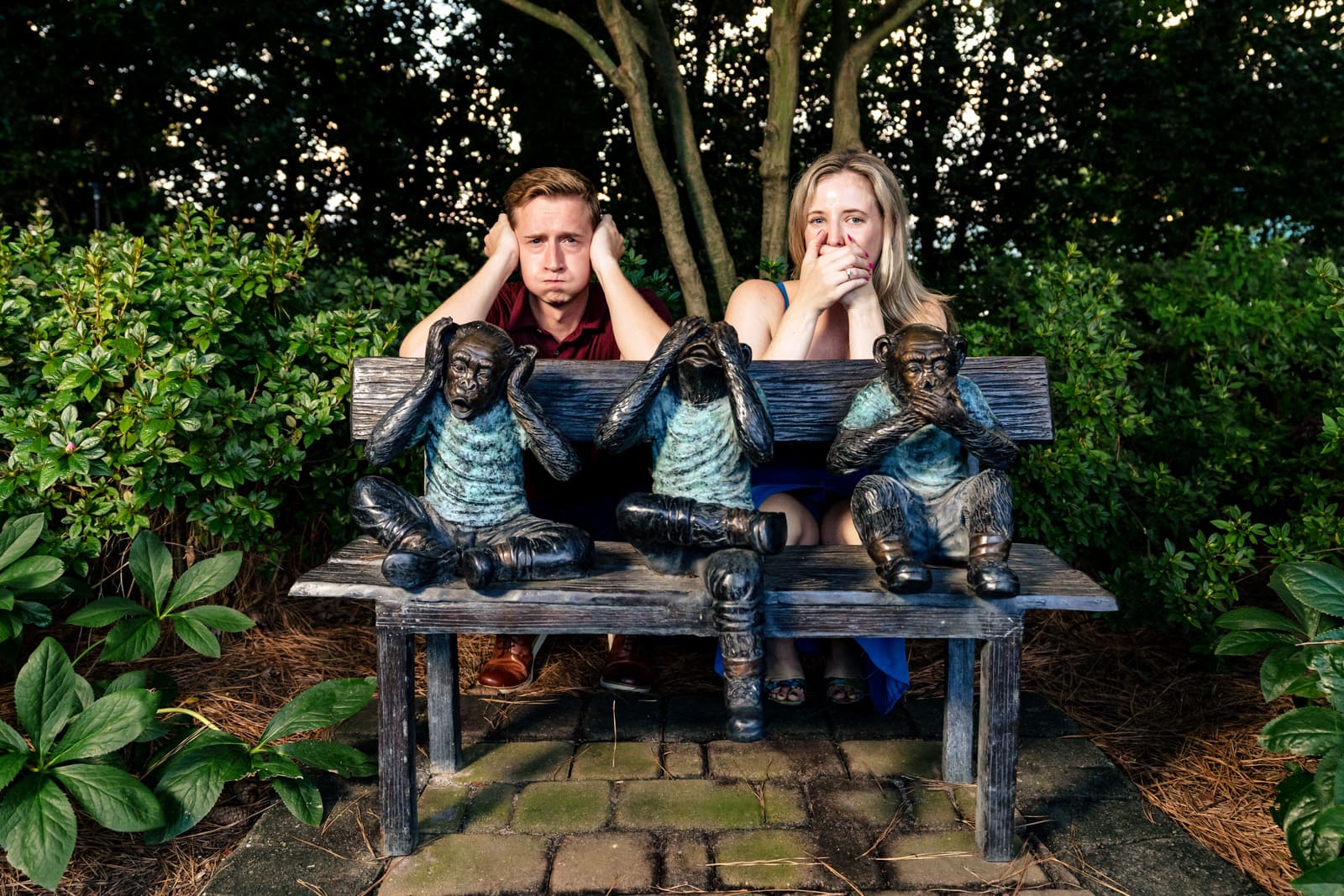 an engaged couple poses behind a sculpture of monkeys on a bench doing the "hear no evil, see no evil, speak no evil pose" creative engagement photography at Killjoy Cocktail bar in Raleigh, NC | photos by Kivus & Camera