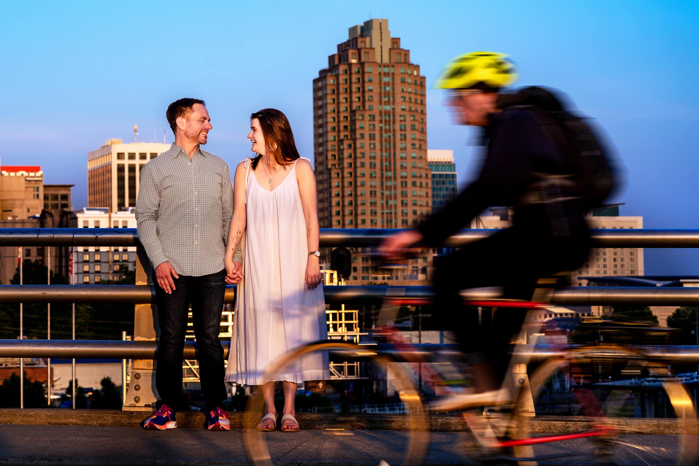 a cyclist passes in front of a couple who are standing on a bridge overlooking the Raleigh skyline