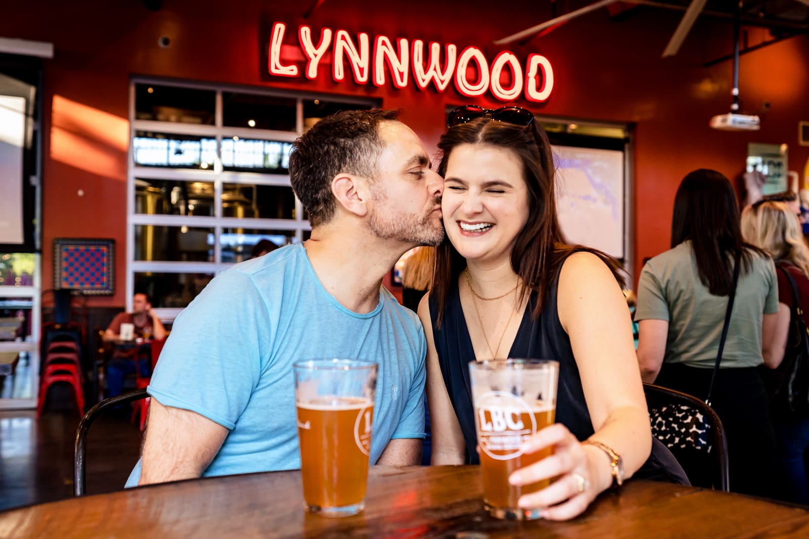 In front a neon sign at Lynwood Brewing Concern in Raleigh, NC a man kisses a woman while she laughs