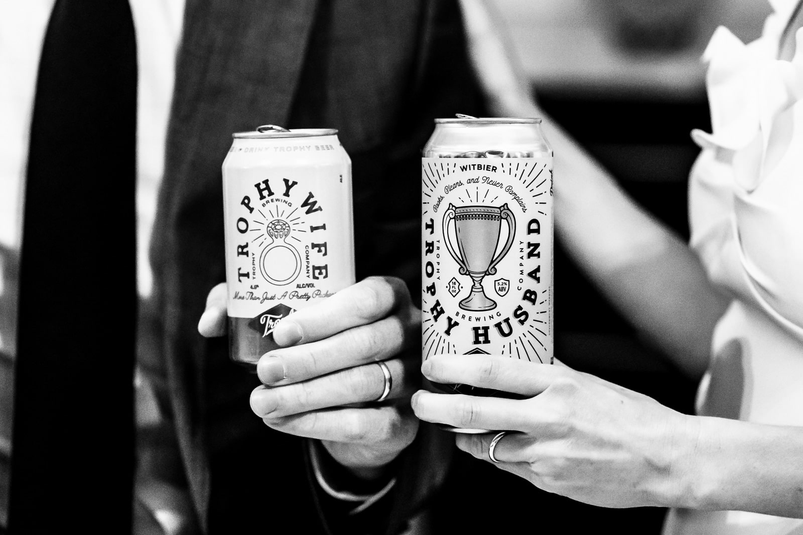 two hands with wedding rings hold beer cans; one says Trophy husband, the other says Trophy wife