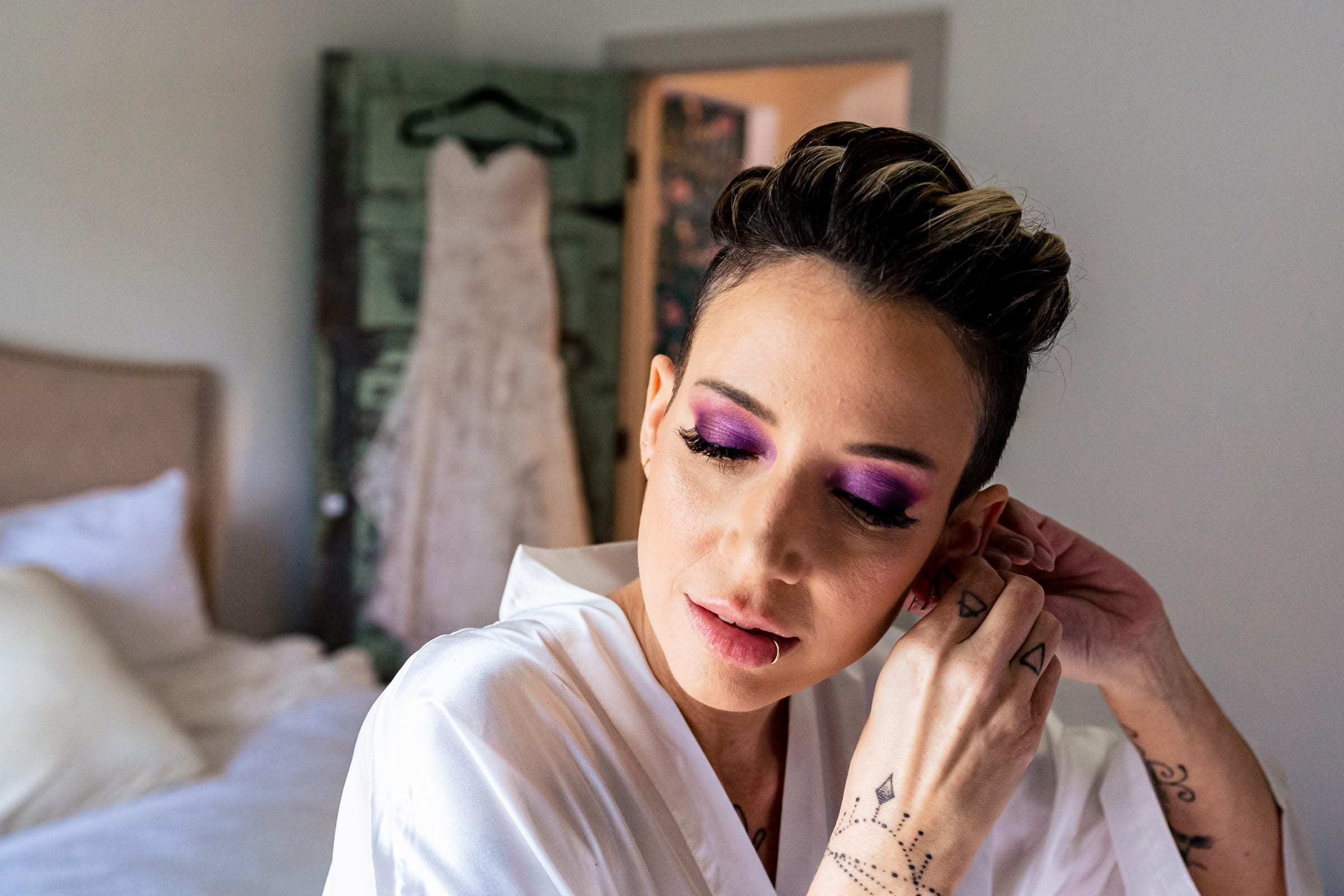 Bride in purple glittery eye shadow puts her earrings in, with her wedding dress visible in the background | Kivus & Camera