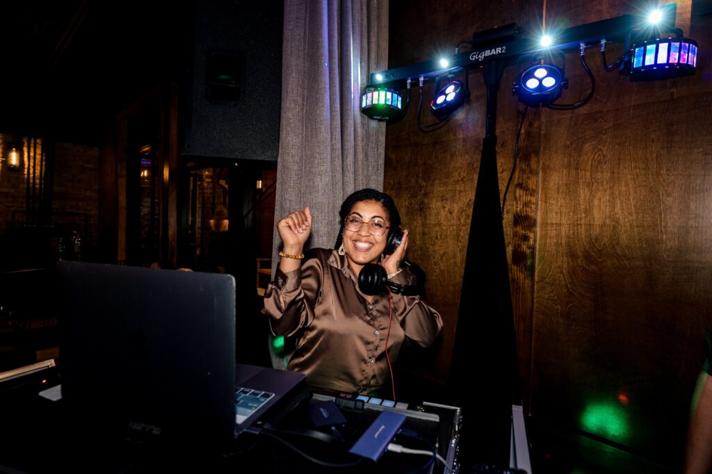 Ava from Raleigh's DJ Ones smiles behind her DJ booth at a wedding | photo by Kivus & Camera