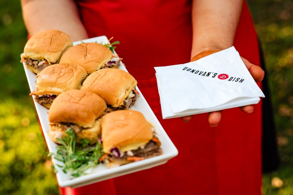 cater-waiter's hands in front of a red apron. one hand is holding a tray of bbq sliders, the other is holding a stack of cocktail napkins. 