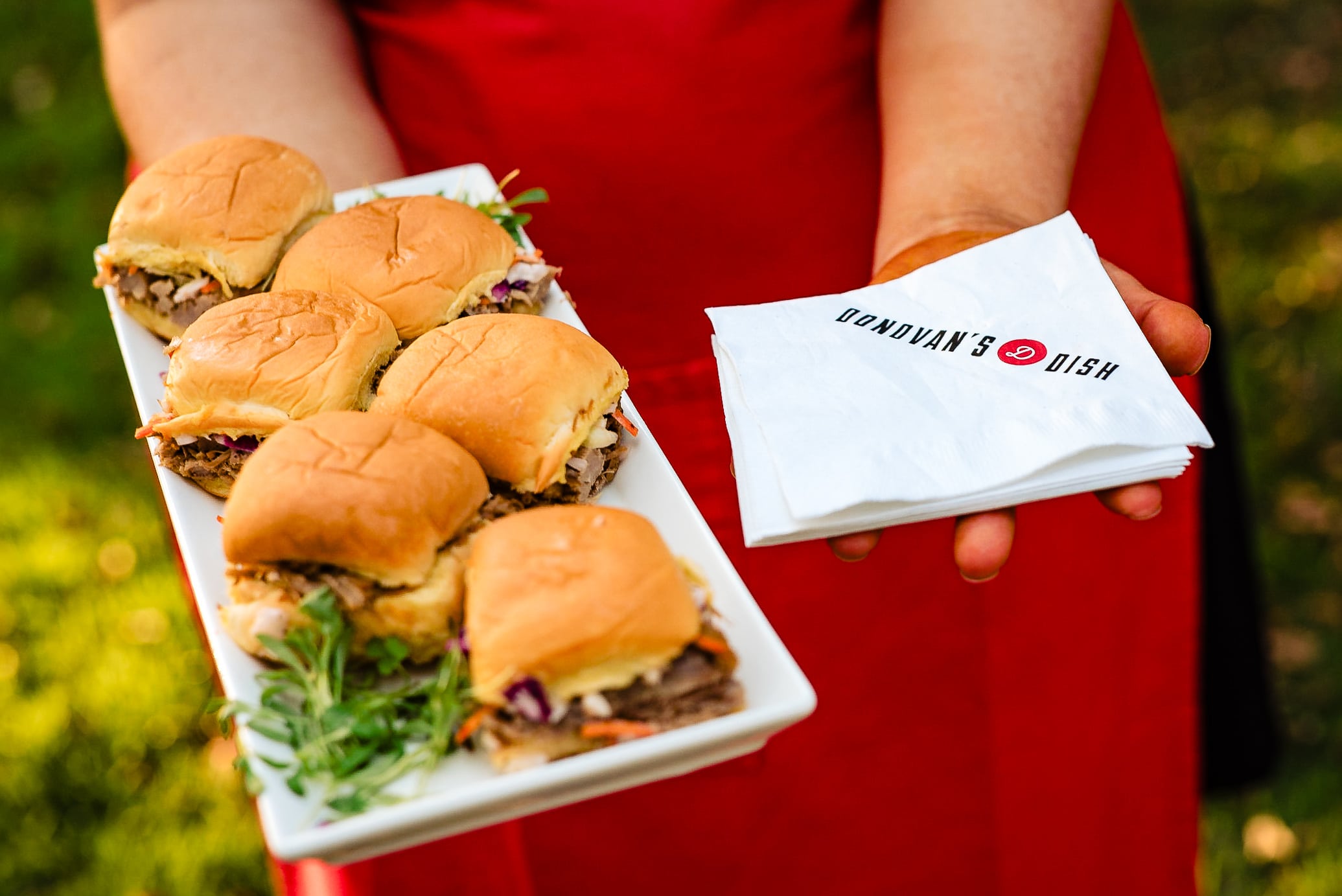 caterer's hands: one holding a tray of bbq sliders, the other holding a stack of cocktail napkins.