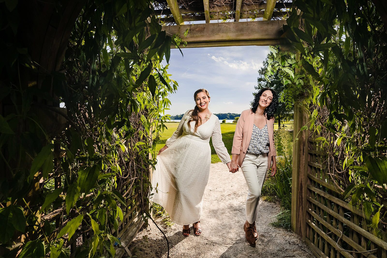 two brides smile on the day of their same-sex elopement in Raleigh, NC. One bride wears a long white dress, the other wears white pants, and a pink blazer.