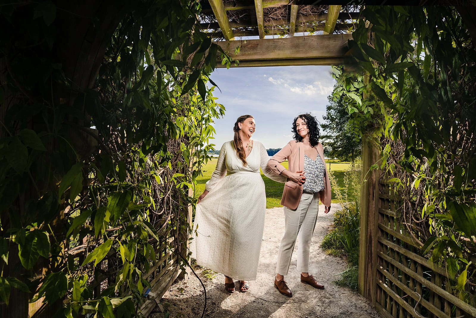 two brides smile on the day of their same-sex elopement in Raleigh, NC. One bride wears a long white dress, the other wears white pants, and a pink blazer.