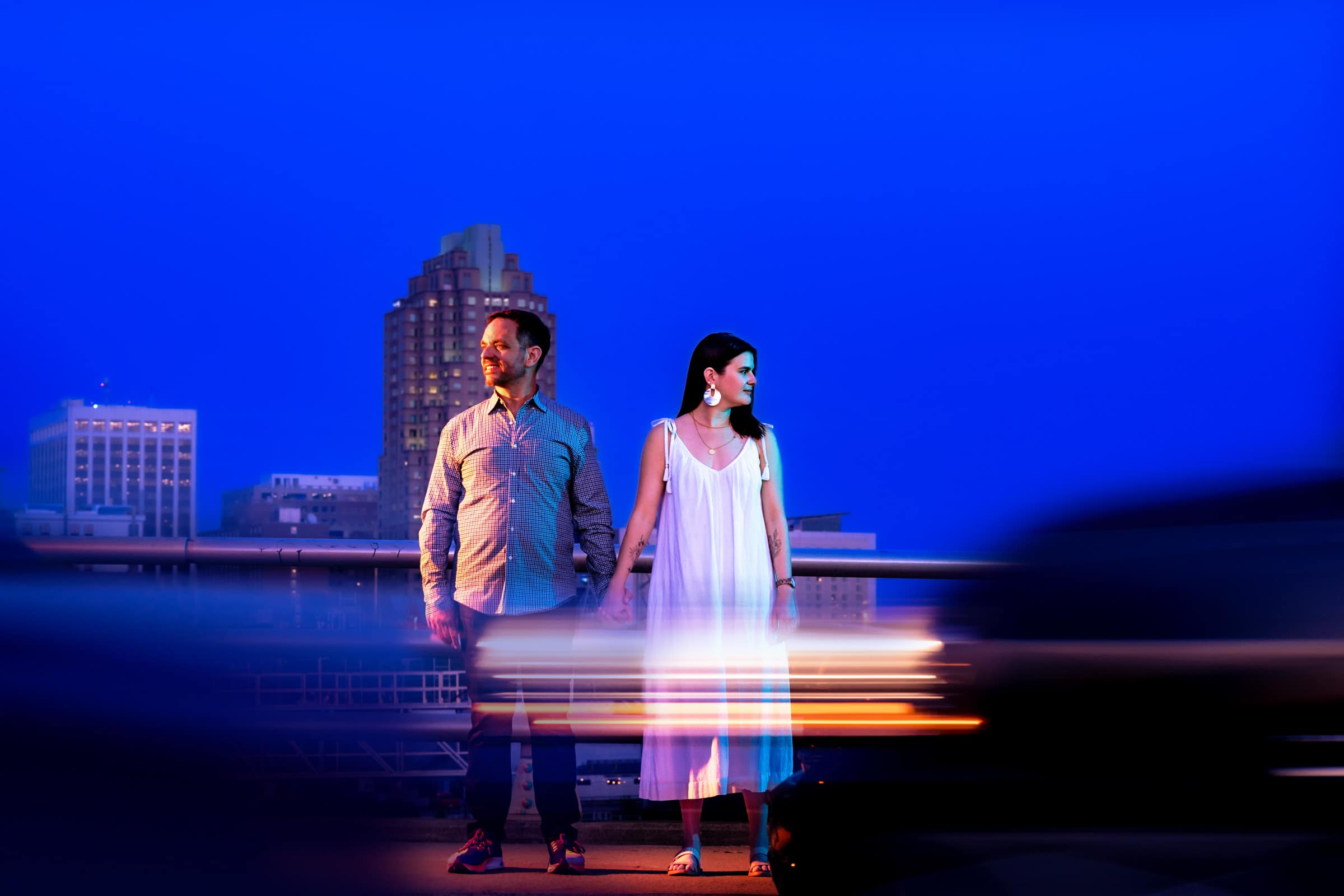 portrait of an engaged couple with the Raleigh skyline in the background behind them, and streaks of car headlights in the foreground | photo by Kivus & Camera