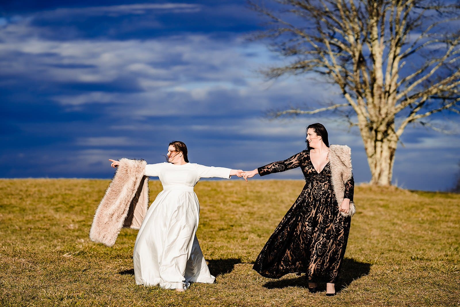 Two brides, one in a black dress and one in a white dress hold hands and walk down a mountain at their elopement in Banner Elk, NC