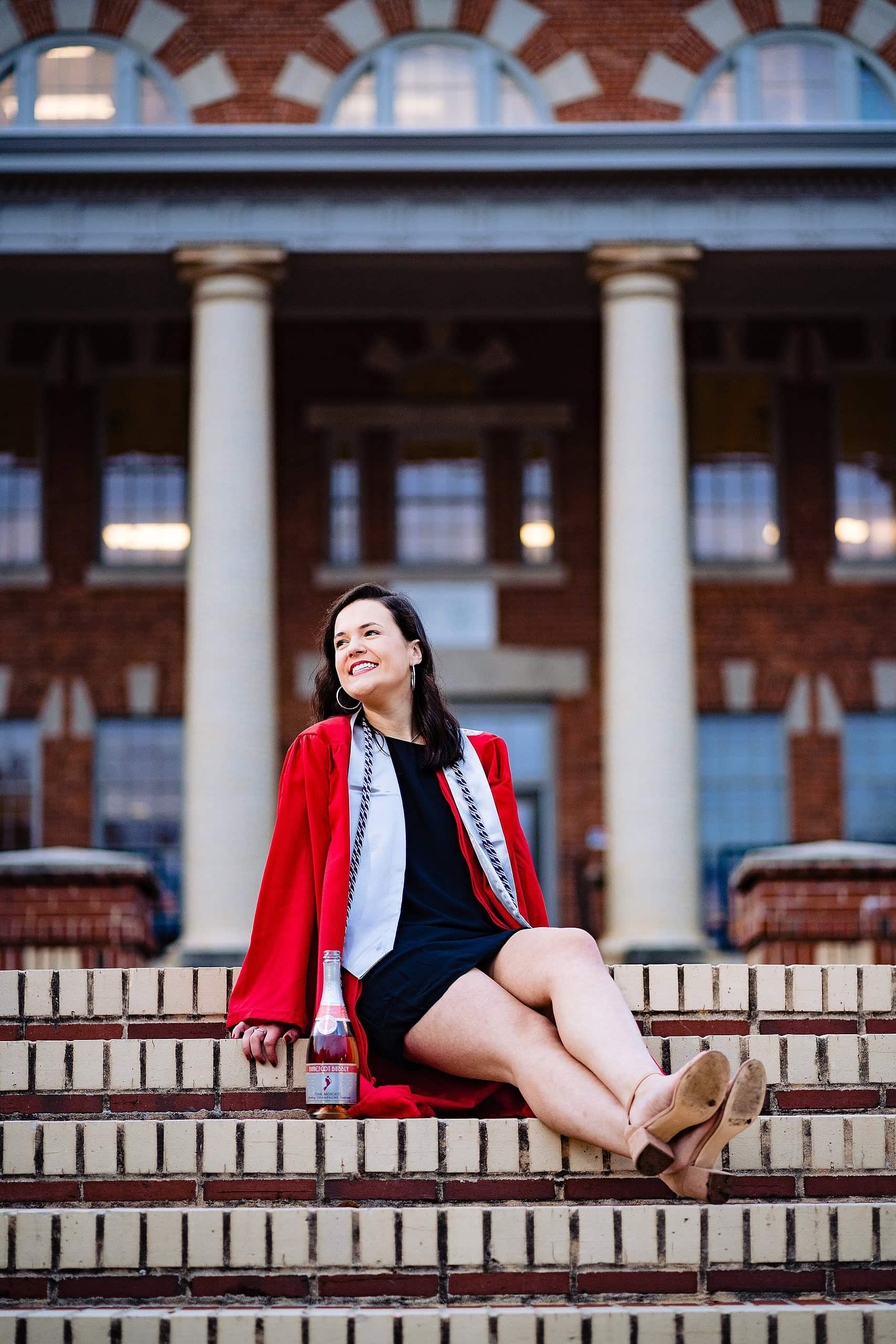 An NC State grad, class of 2019, smiles during graduation portraits. She's wearing her red robe and sitting at the top of the steps in front of the 1911 Building bordering the Court of Carolinas | kivusandcamera.com