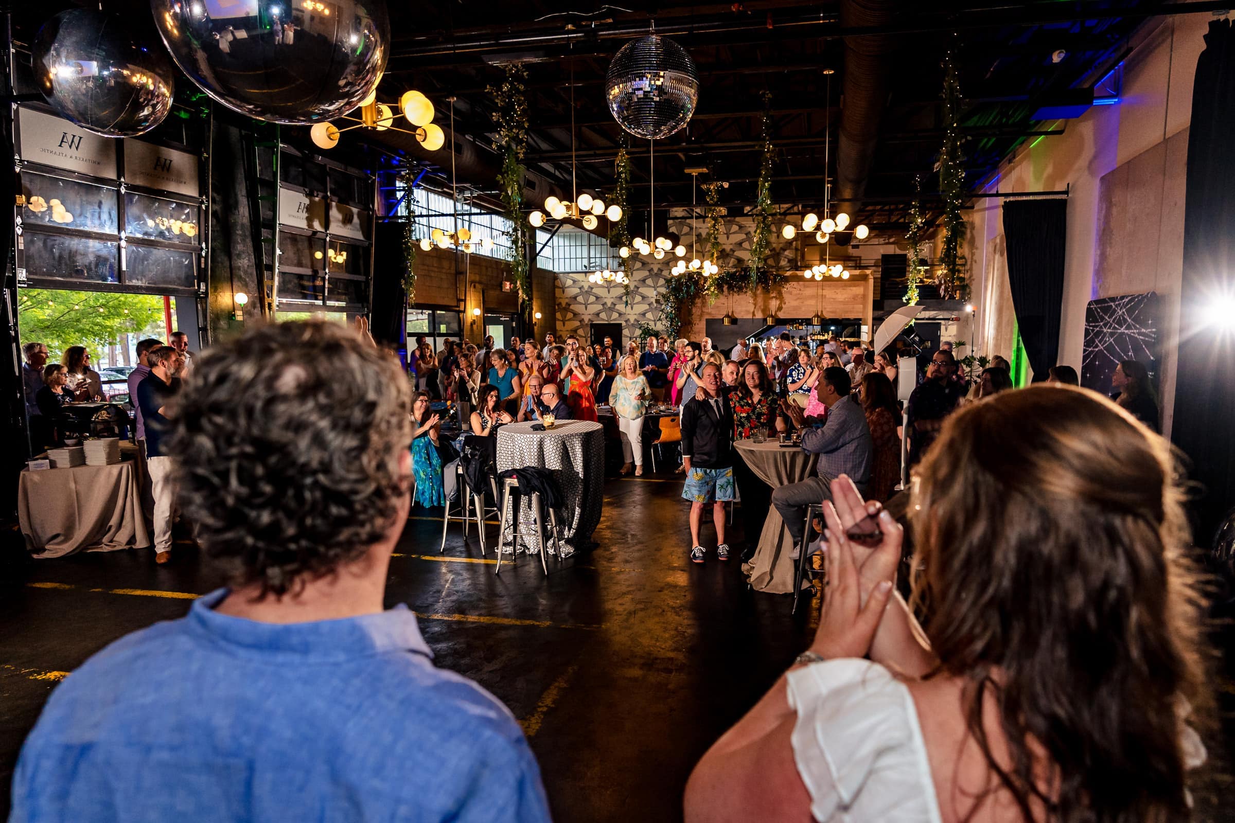 Wedding guests stand and applaud with the couple visible facing the guests in the foreground