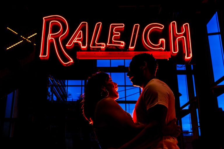 a couple is back lit by a red neon sign that says 'Raleigh'; behind them the sky is vivid blue during blue hour after the sunset