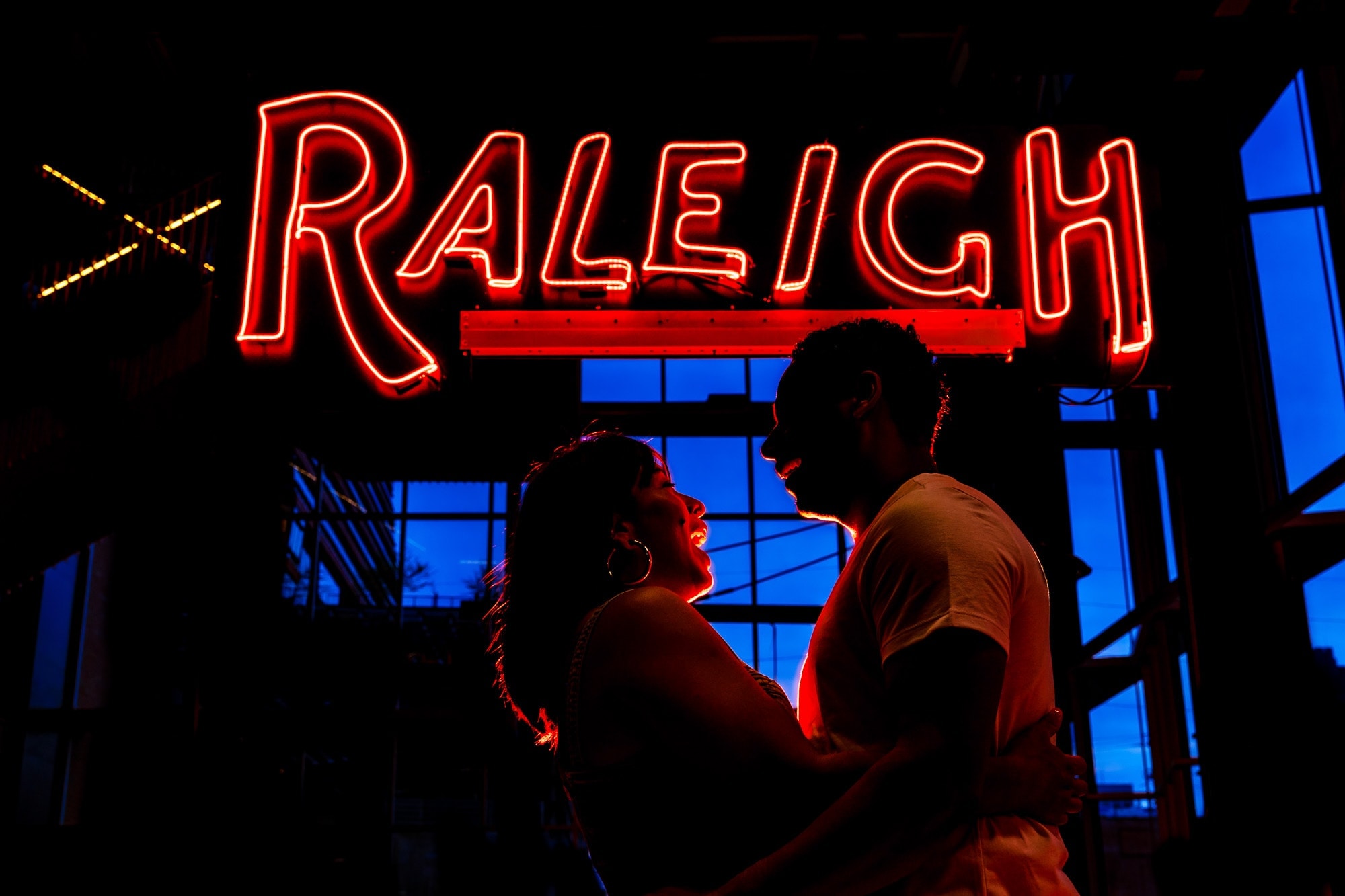 a couple is back lit by a red neon sign that says 'Raleigh'; behind them the sky is vivid blue during blue hour after the sunset