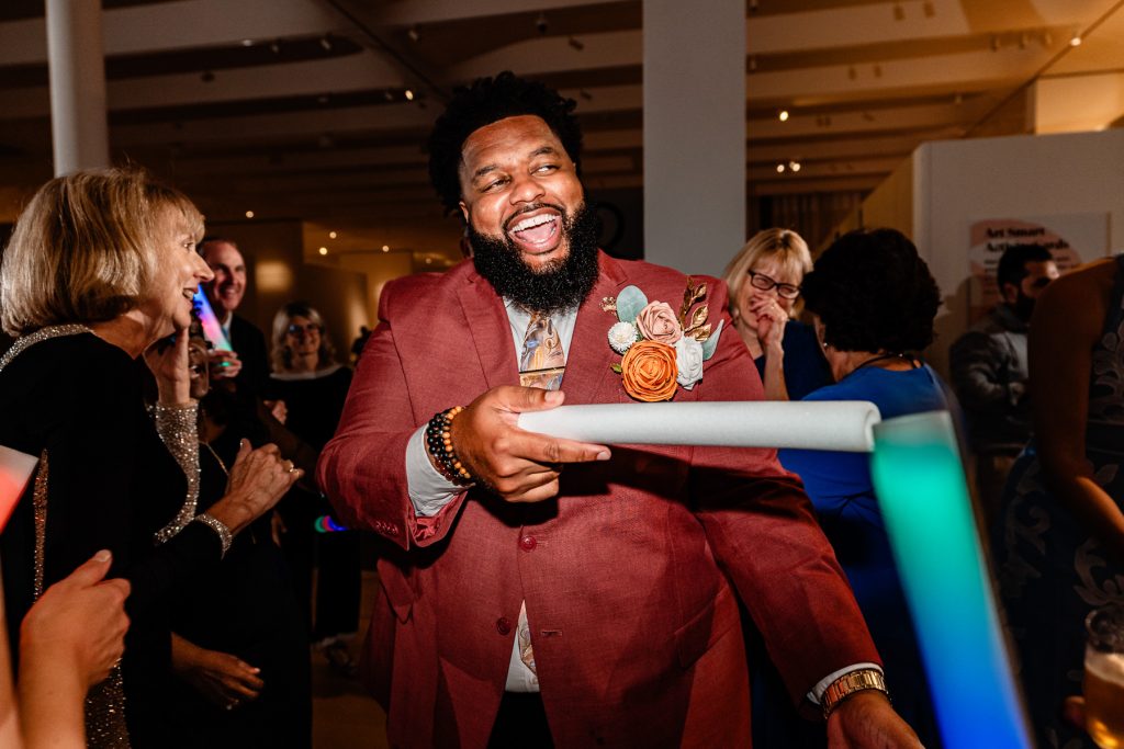 man in formal attire smiles and waves a glow stick as he dances at a wedding