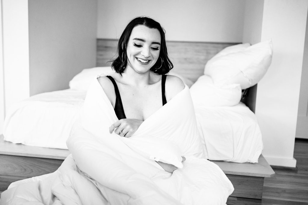 photo of a woman laughing sitting at the foot of a bed with a comforter wrapped around her | photo by Triangle Boudoir