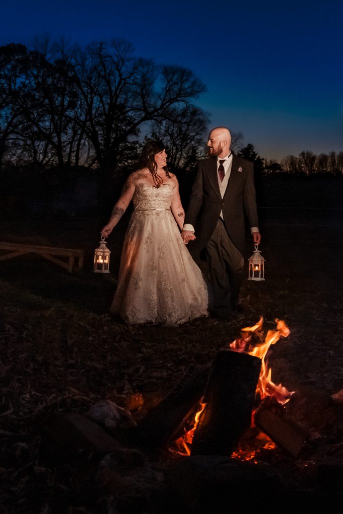 couple in wedding attire walk through the dark towards a fire pit while holding lanterns