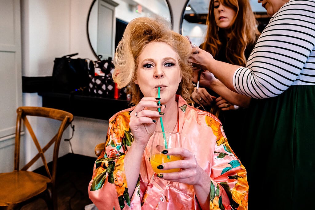 bride sips on a mimosa as her hair and makeup are being done