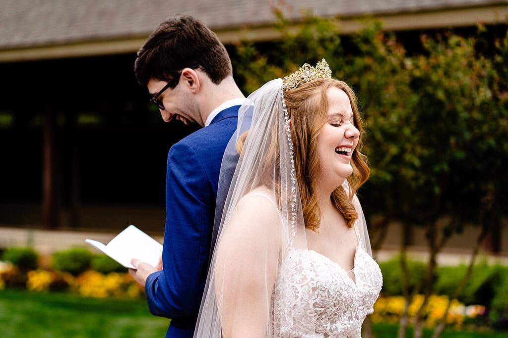 bride and groom stand back to back. bride is laughing at something the groom is reading from a vow book.