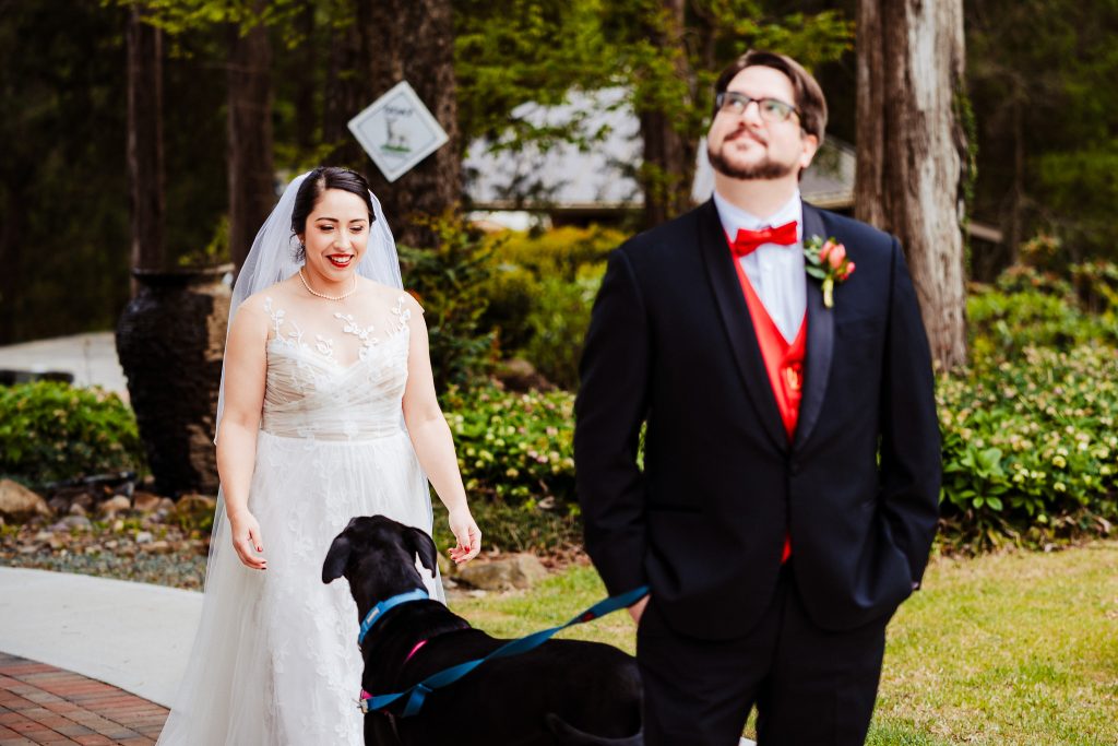 man in a tuxedo looks into the distance, the dog next to him is looking at a woman in a wedding dress who is approaching the man from behind for the couple to share a first look on their wedding day | photo by Kivus & Camera