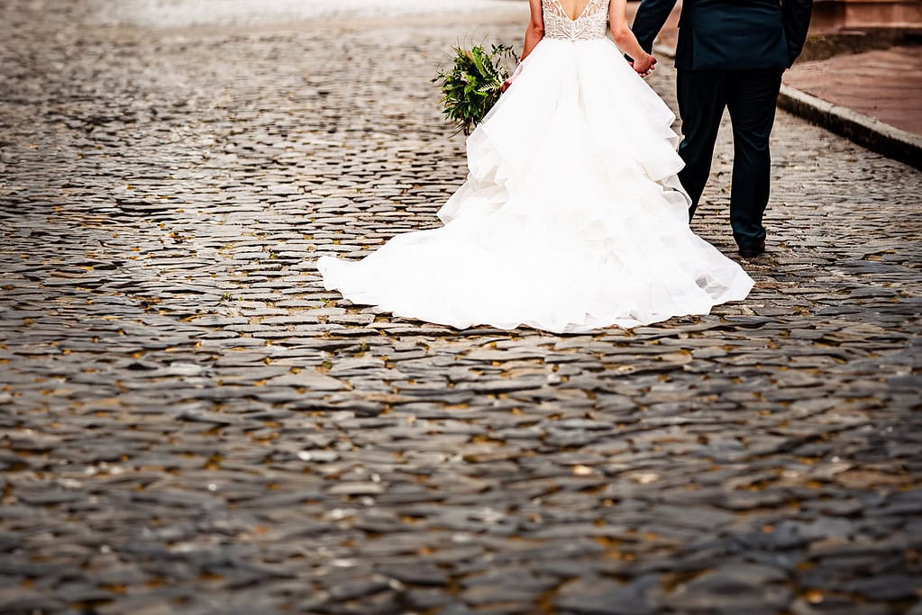 a couple in wedding attire holds hands as they walk away from the camera down a cobblestone street
