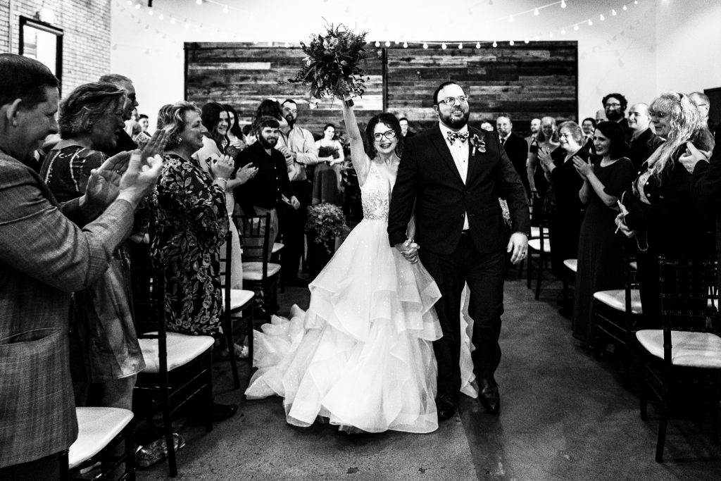 bride and groom processing back down the aisle as their guests applaud and bride holds bouquet in the air in celebration