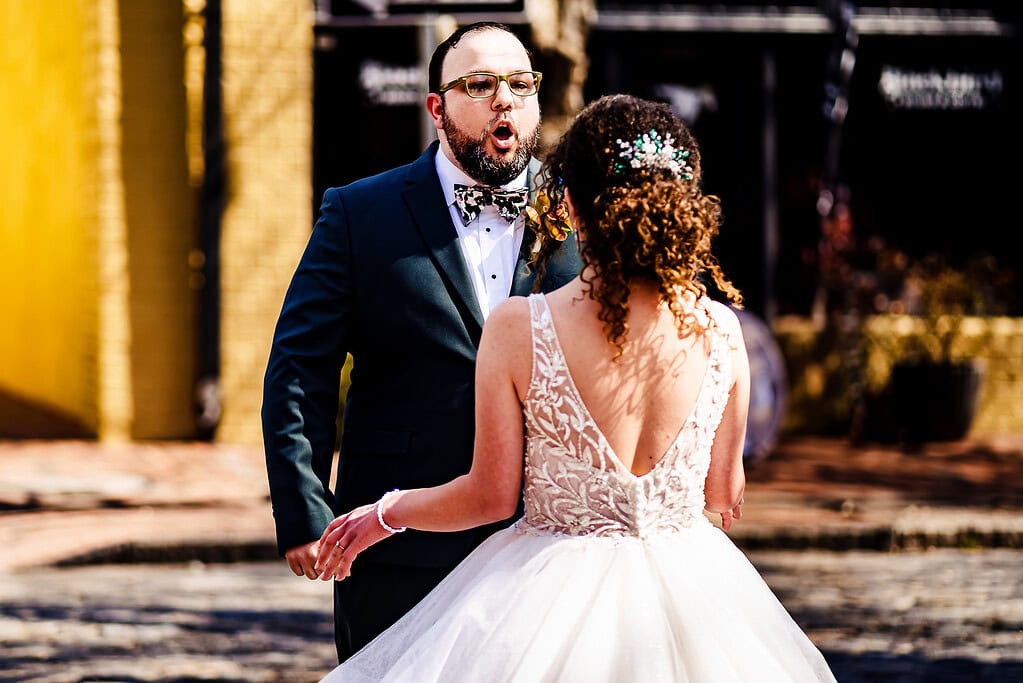 groom in a green suit and floral bowtie reacts with happiness as he sees bride for the first time on their wedding day