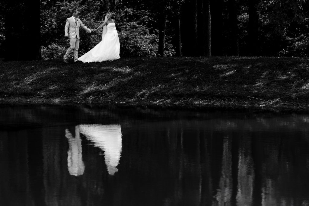 black and white portrait of a bride and groom walking alongside a lake. their reflections appear below.