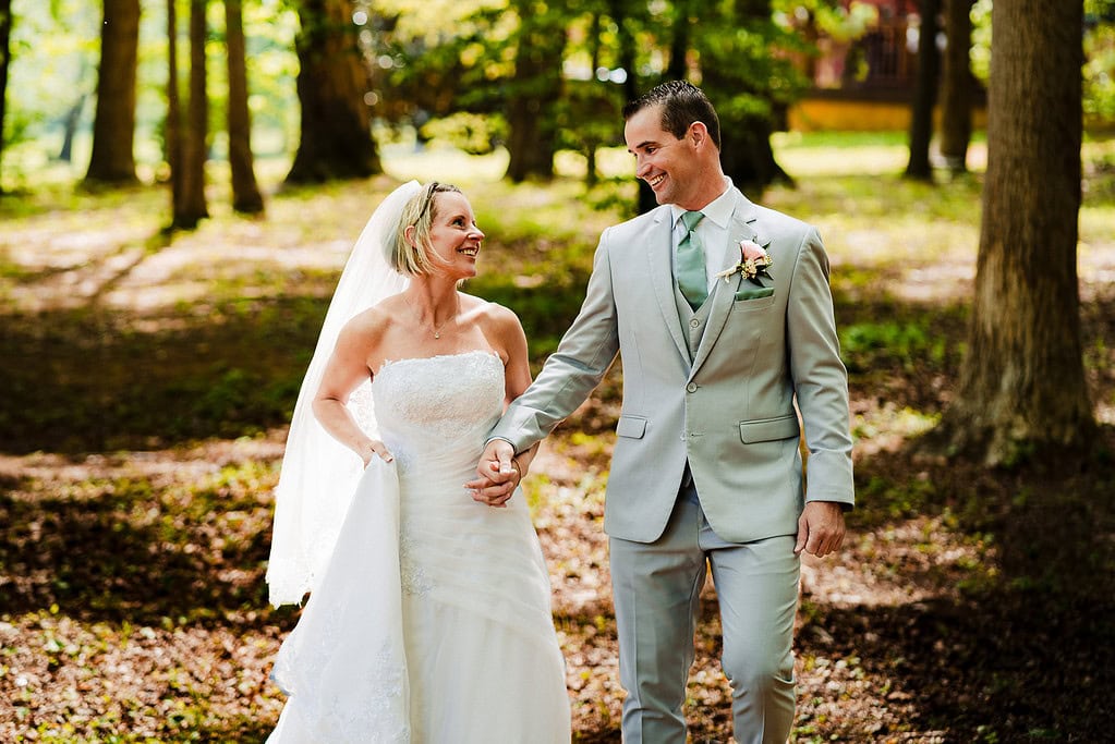 couple in wedding attire walks hand in hand through a treed area 