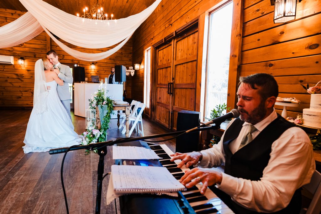 man in formal attire sings and plays piano, while in the background a bride and groom are dancing