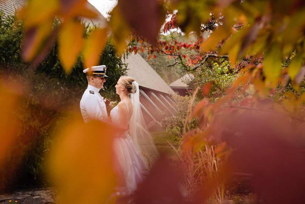 Woman in a wedding dress embraces a man in military dress whites on their wedding day. The couple are framed amongst autumn leaves | photo by Kivus & Camera