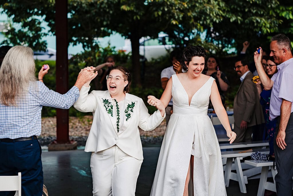 Two women, one in a white suit, one in a white dress hold hands and smile as the exit their wedding ceremony | photo by Kivus & Camera