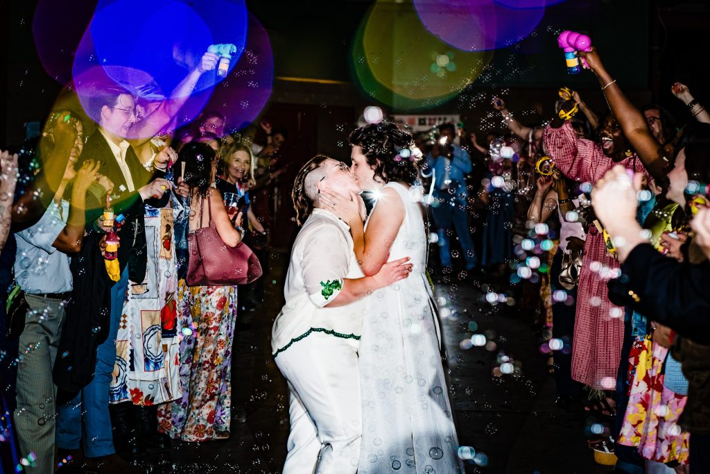 two women dressed in white bridal attire share a kiss while surrounded by guests blowing bubbles | photo by Kivus & Camera